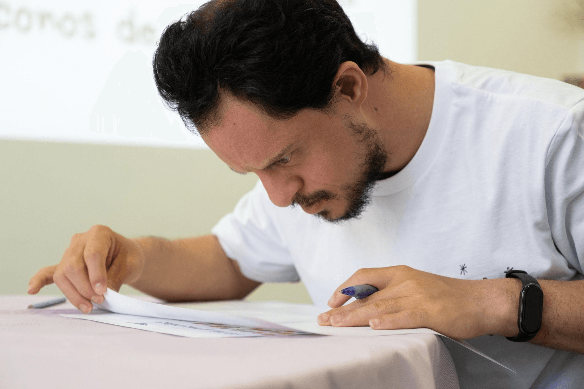 Hombre concentrado revisando un documento en una mesa, sosteniendo un bolígrafo y vistiendo una camiseta blanca, en un entorno de trabajo tranquilo.