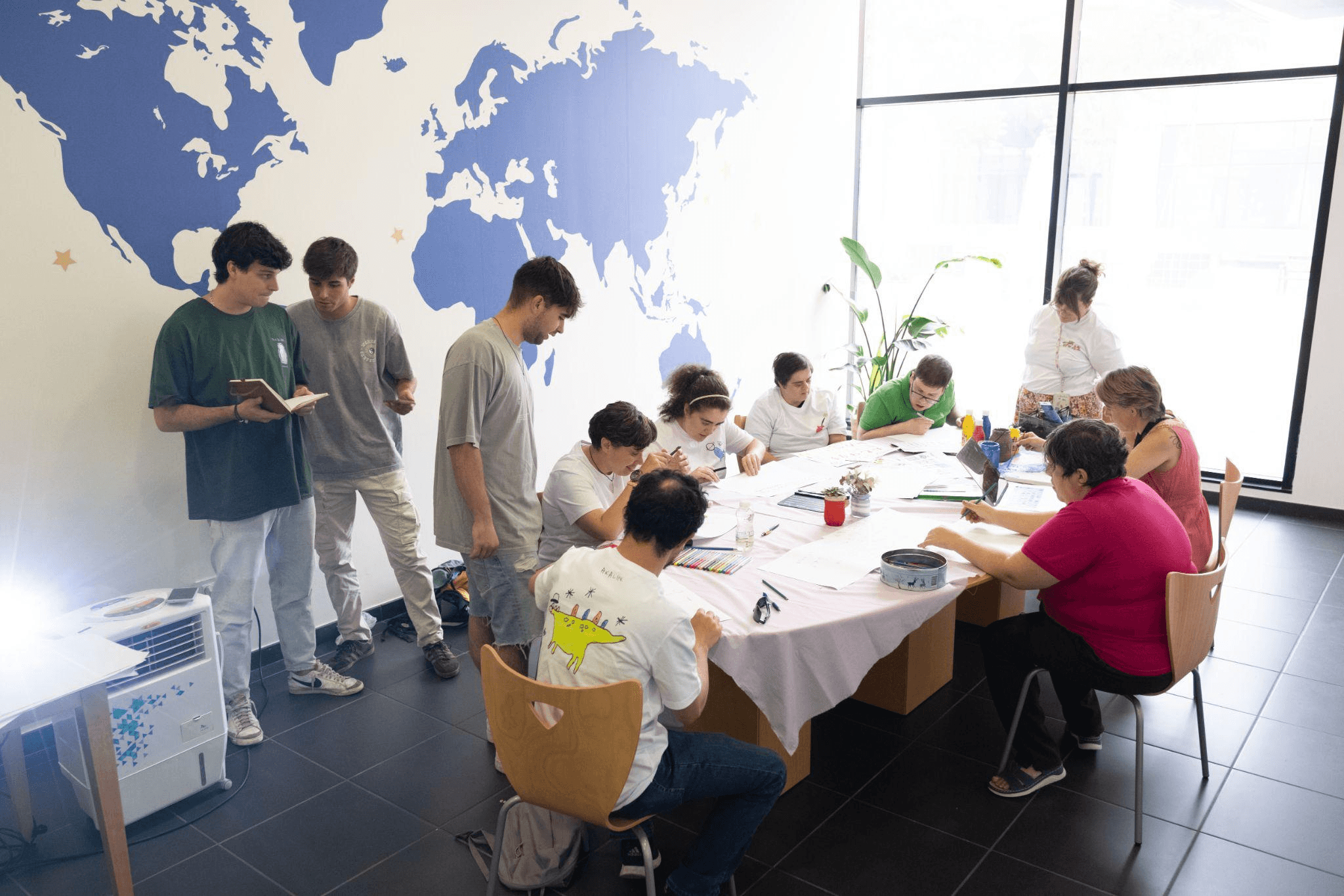 Grupo de personas trabajando en una actividad creativa alrededor de una mesa en una sala iluminada, decorada con un mapa mundial en la pared, mientras algunos observan y colaboran.