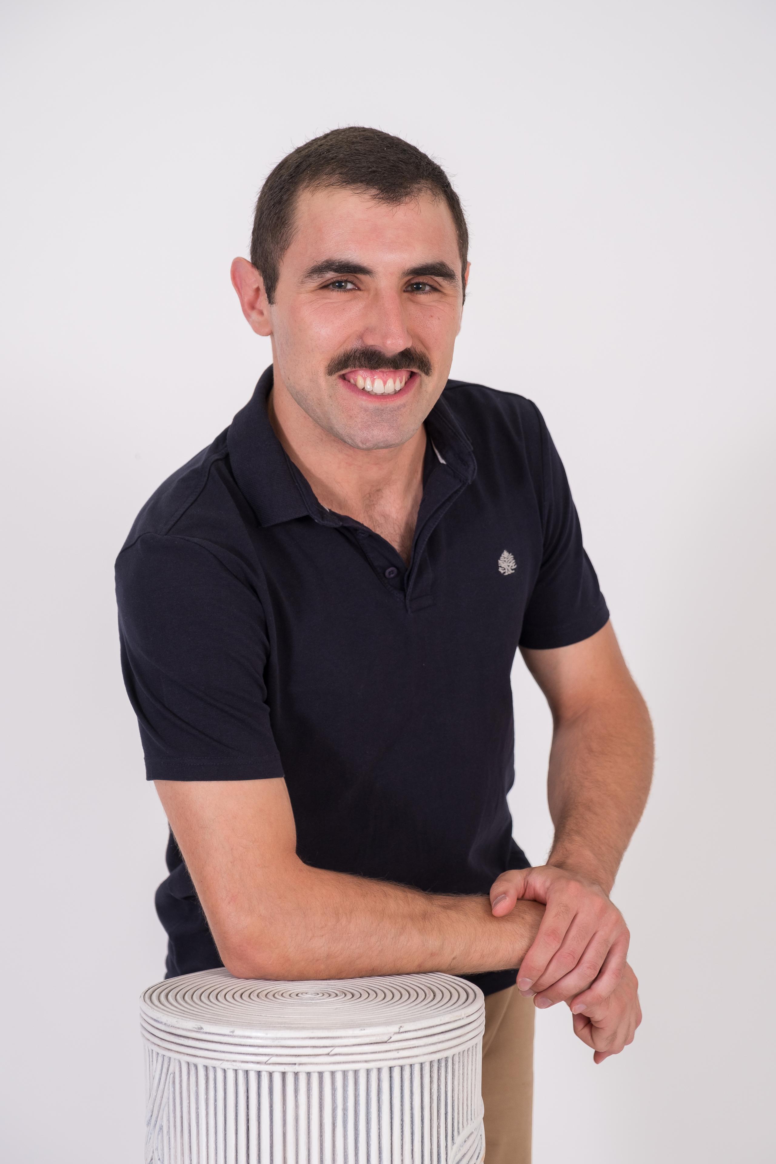 Hombre sonriente con polo negro posando junto a un cilindro decorativo en fondo blanco.