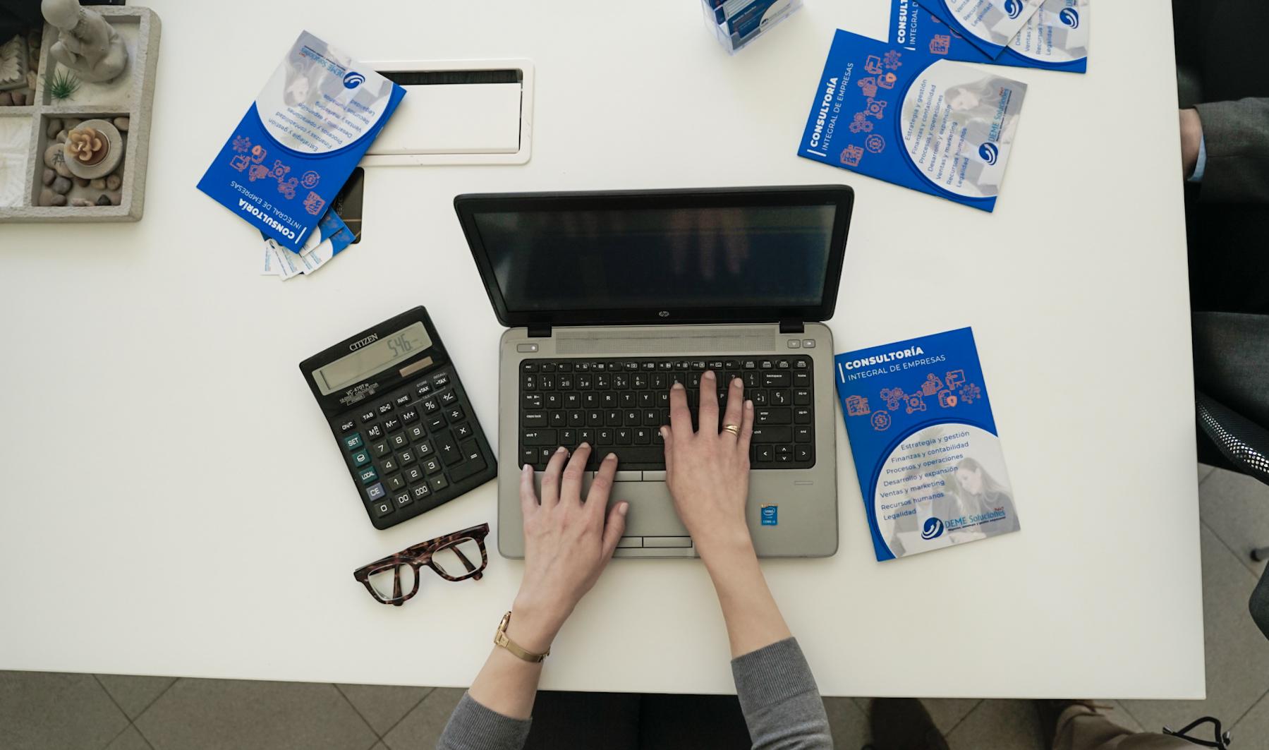Persona trabajando en una computadora portátil sobre un escritorio blanco, rodeada de folletos de consultoría empresarial, una calculadora y unas gafas de lectura.