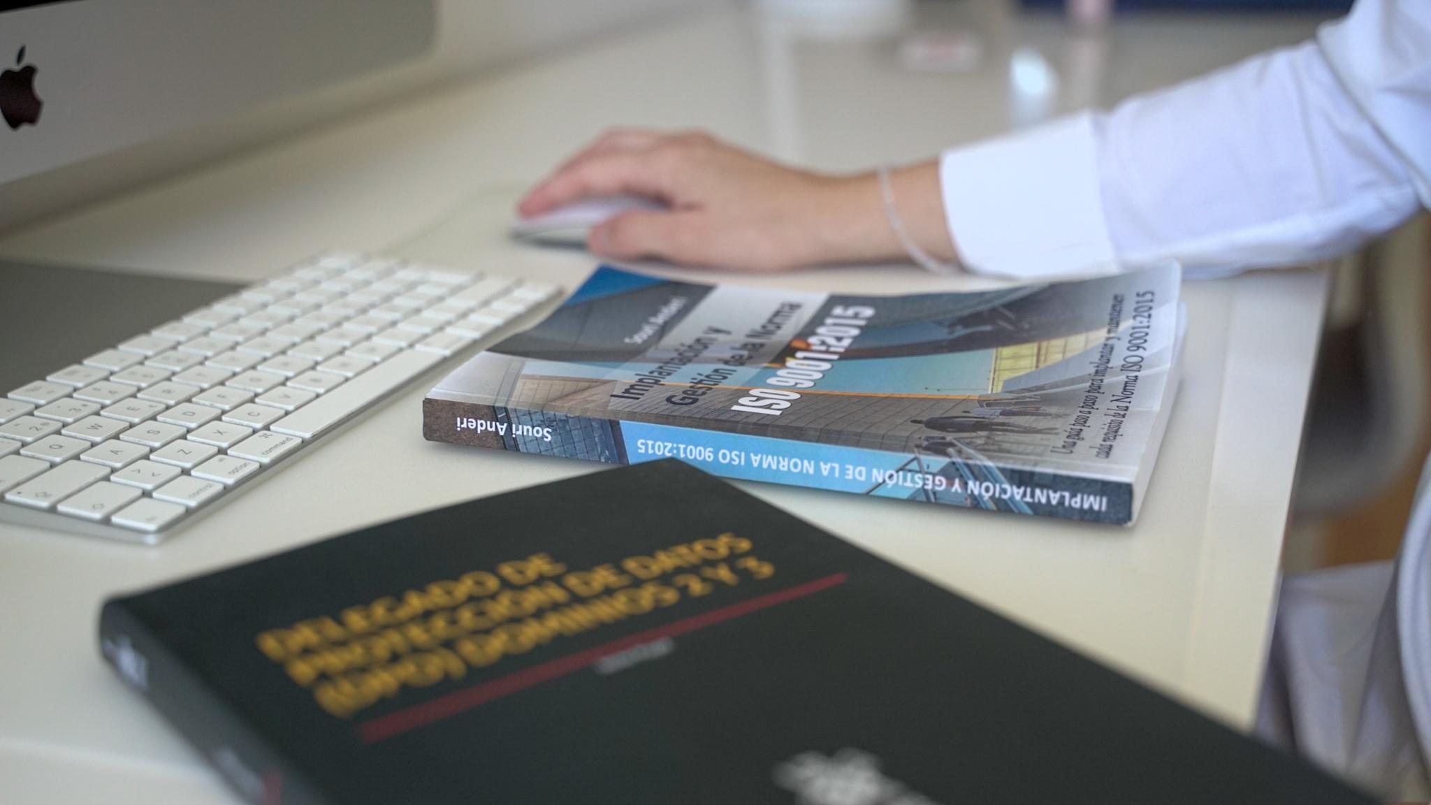 Persona utilizando un ratón junto a un teclado en un escritorio, con libros sobre gestión y normas ISO.