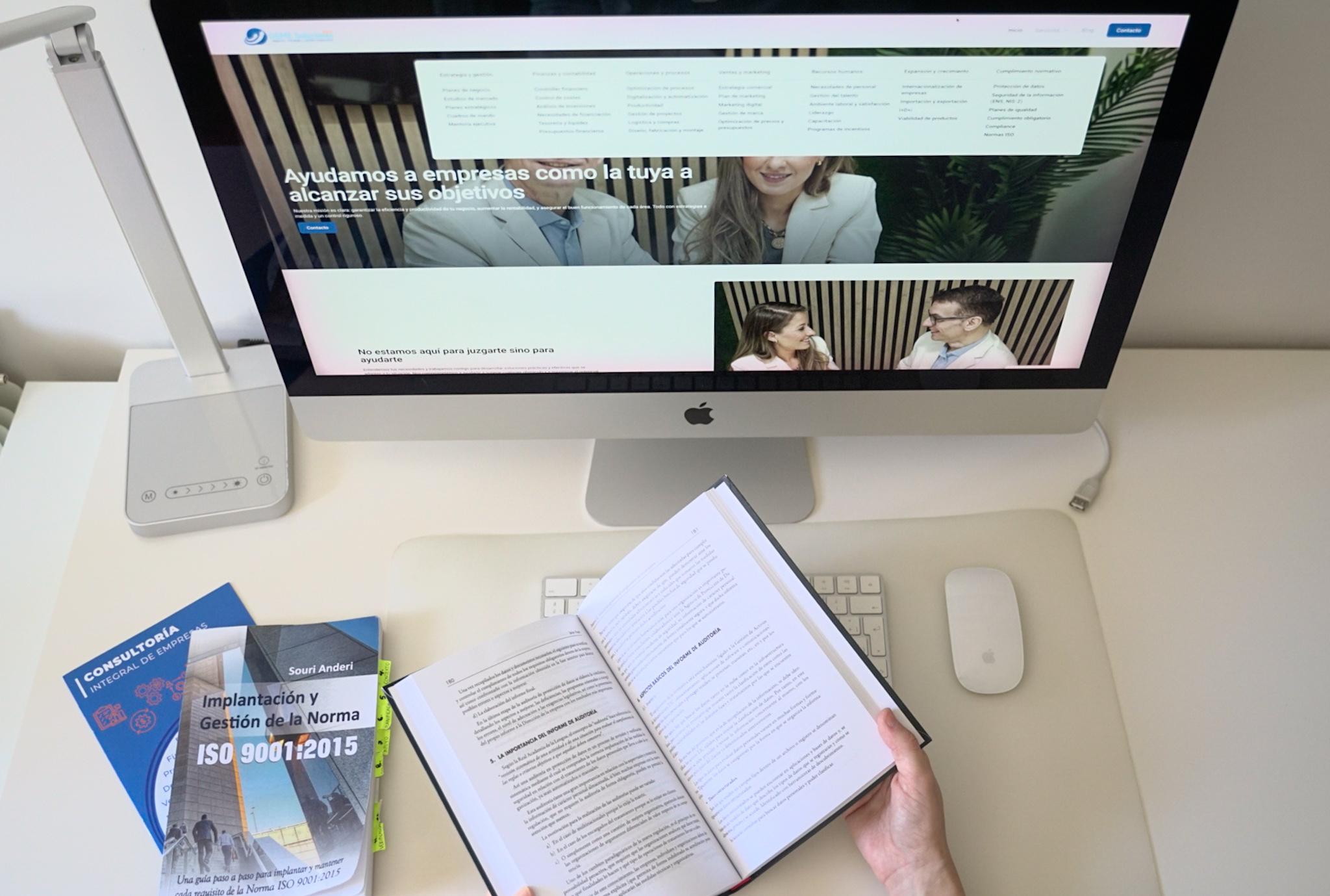 Persona leyendo un libro sobre normas ISO frente a una computadora con un sitio web de consultoría empresarial en la pantalla.