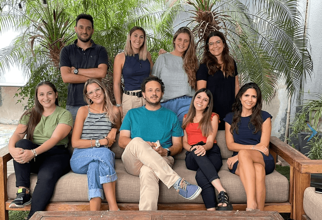 Grupo de personas posa para una foto rodeado de plantas tropicales. Están sentados y de pie en un sofá de madera, con expresiones relajadas y sonrientes. La escena tiene un aire casual y natural, con todos vestidos en ropa informal y cómoda.