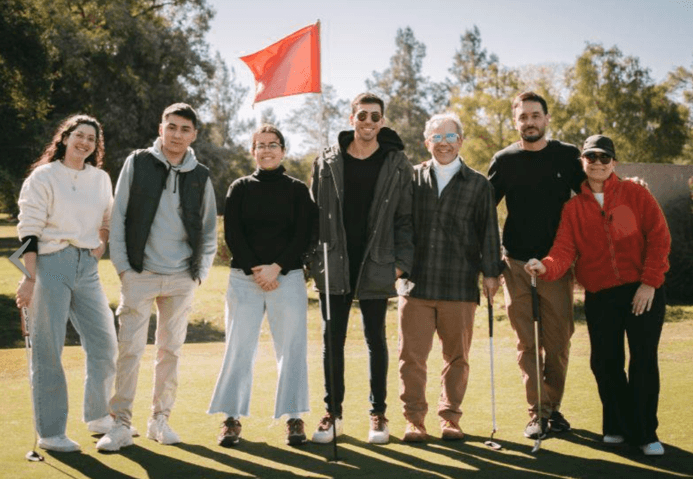 Grupo de personas posa en un campo de golf al aire libre bajo un cielo despejado. Están de pie junto a una bandera roja que marca un hoyo en el campo. Visten ropa casual y abrigada; algunos llevan gafas de sol y chaquetas. El ambiente es relajado, con sonrisas y una postura amigable entre los integrantes. Detrás, se observan árboles y un paisaje natural, resaltando el clima soleado y la vegetación verde.