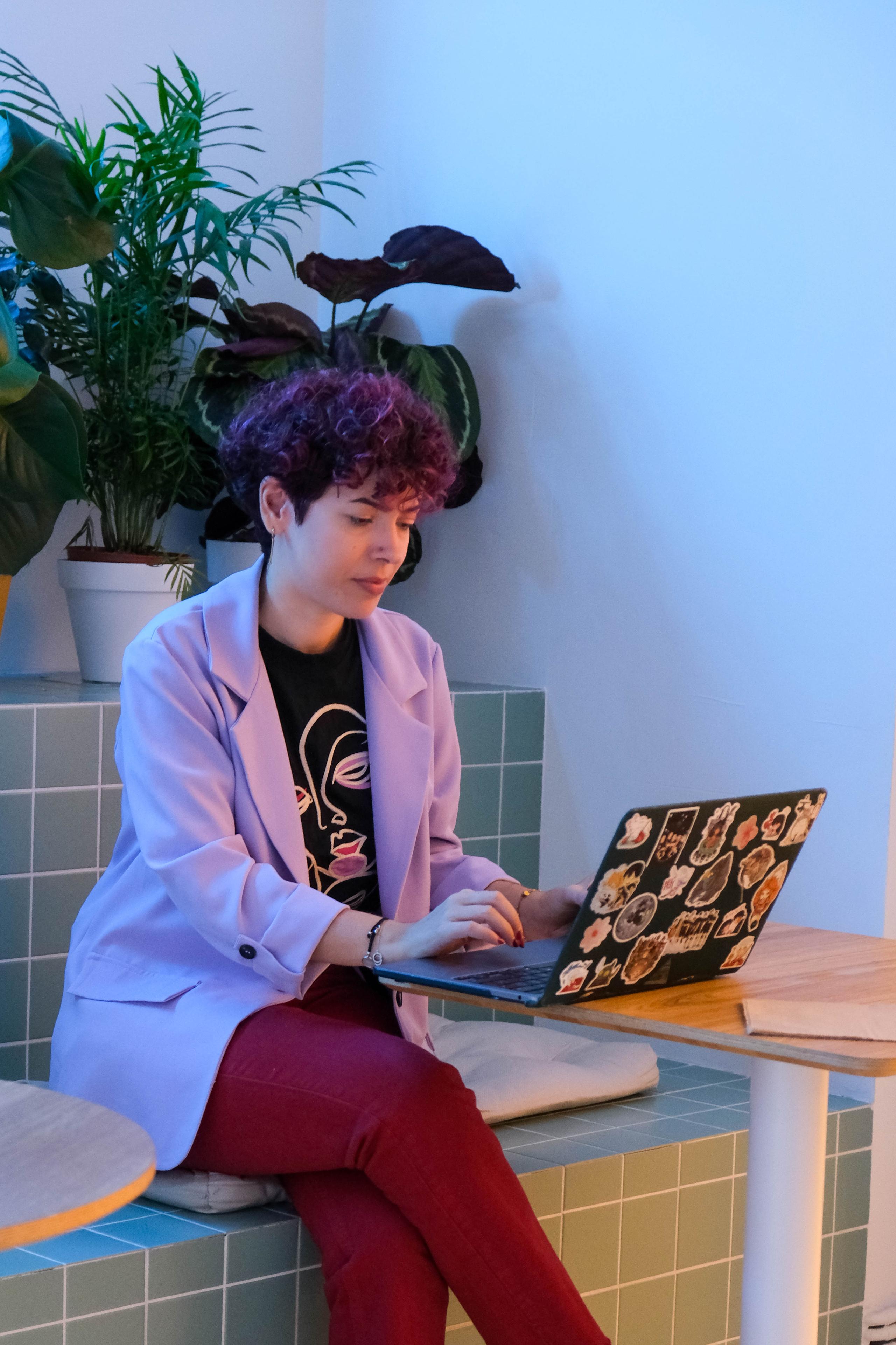 Persona trabajando en un portátil decorado con pegatinas, rodeada de plantas en un espacio moderno.