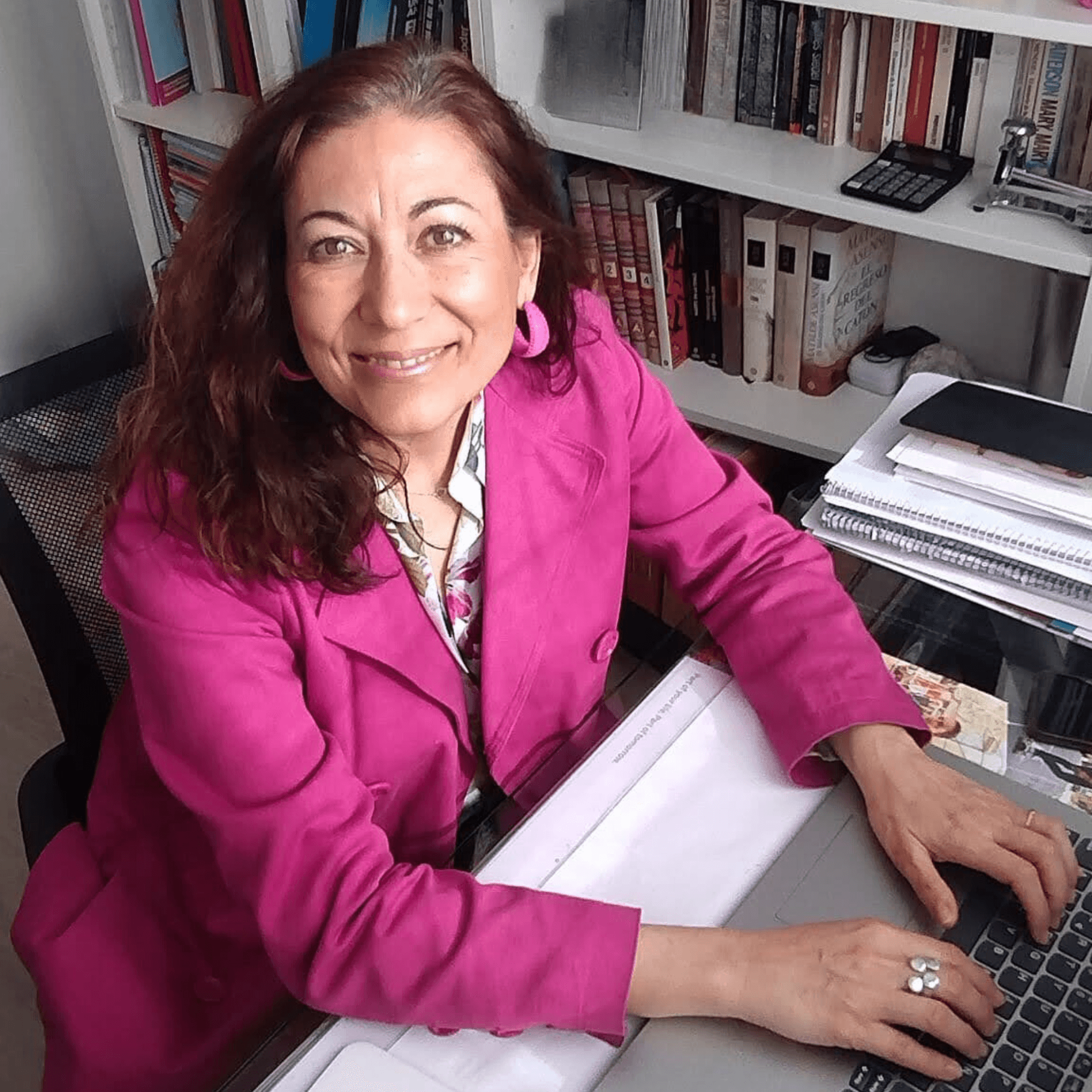 Mujer sonriente sentada frente a un escritorio, usando un ordenador portátil. Lleva una chaqueta de color fucsia y pendientes grandes a juego. Detrás de ella hay una estantería llena de libros y algunos objetos de oficina, creando un ambiente acogedor y profesional. Su expresión es cálida y amigable, y parece estar trabajando.